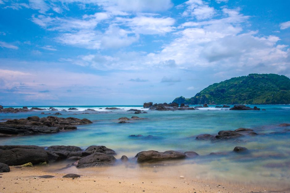 Idyllic tropical beach scene with rocky shoreline, clear blue waters, and lush hill in the background.