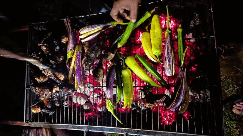 Grilled eggplants, peppers, and chicken wings over glowing coconut shell embers at night